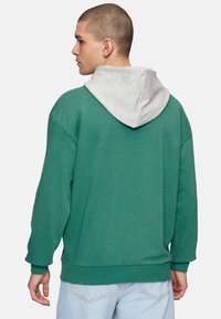 Felpa verde con cappuccio grigio, vestibilità oversize, spalle cadute e polsini a coste. Il tessuto sembra morbido e informale.