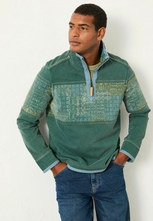 Grüner Fleece-Pullover mit halbem Reißverschluss-Kragen, gemustertem Brustpanel und kontrastierendem blauen Besatz. Auffällige Texturen und Stickdetails.
