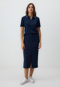 Polo shirt blu navy con maniche corte e colletto con zip, abbinato a una gonna al ginocchio con spacco frontale, indossato con sneakers bianche.