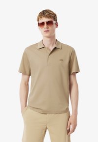 Beige pikétröja med klassisk krage och korta ärmar, med en liten logotyp på bröstet. Materialet har en slät textur.