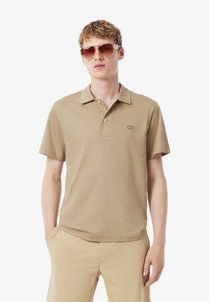 Polo beige con colletto classico e maniche corte, dotato di un piccolo logo sul petto. Il materiale ha una texture liscia.