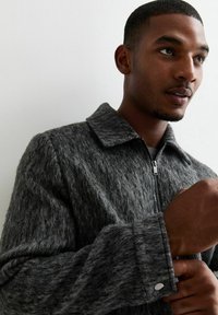 Chaqueta gris texturizada con cremallera y cuello tipo solapa, de mangas largas, que presenta una cremallera plateada y un detalle de broche en el puño.