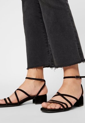 Fødder iført sorte sandaler med lav hæl og stropper, kombineret med korte sorte jeans med frynset kant mod en ensfarvet baggrund.