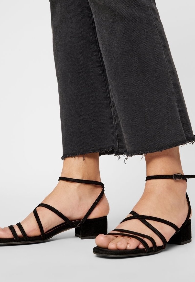 Pieds portant des sandales noires à brides et talons bas, associées à un jean noir court avec des ourlets effilochés sur un fond uni.