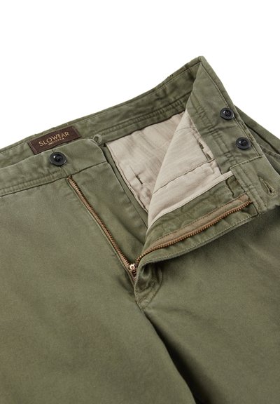 Spodnie z bawełny w oliwkowym zielonym kolorze z zamkiem błyskawicznym, podwójnymi guzikami i beżową podszewką. Charakteryzują się delikatną fakturą i kieszeniami wewnętrznymi.