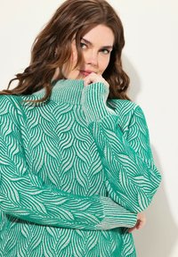 Pull en tricot vert avec un col montant et un motif en vagues. Présente des poignets côtelés et une texture contrastant des fils doux et fermes.