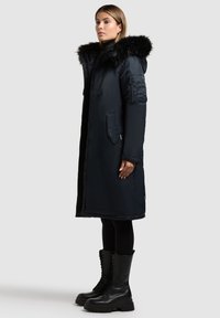 Parka nera lunga con cappuccio in pelliccia sintetica, tasche con zip e dettagli trapuntati sulle maniche; abbinata a stivali militari neri con lacci.