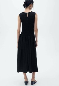 Abito lungo nero con corpetto smock, dettaglio a forma di chiave sulla schiena e parte inferiore fluida, abbinato a sandali con plateau. Texture liscia e design elegante.