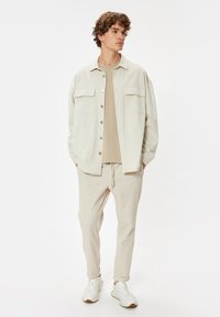 Chaqueta beige claro con dos bolsillos en el pecho, combinada con una camisa beige a juego y pantalones de color claro. Llevada con zapatillas blancas.