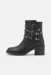 Botas de tobillo de cuero negro con tres correas decorativas adornadas con tachuelas plateadas y detalles de hebilla, tacón robusto y textura suave.