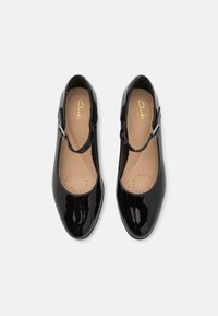 Une paire de chaussures Mary Jane en cuir verni noir avec des semelles intérieures rembourrées beiges, vue de dessus sur un fond blanc.