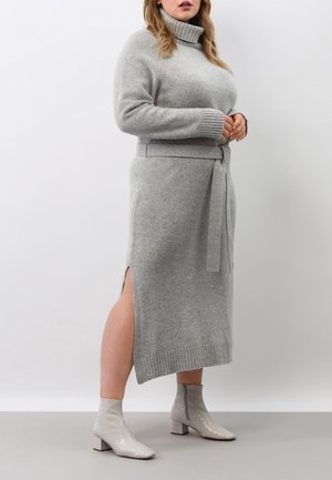 Vrouw draagt een grijze gebreide coljurk met zijsplit en bijpassende riem, gecombineerd met lichtgrijze enkellaarsjes, staand tegen een witte achtergrond.