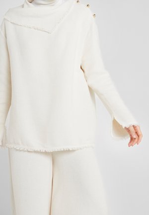 Vrouw draagt een crèmekleurige gebreide trui met gouden knopen op de schouder en bijpassende wijde broek tegen een effen achtergrond.