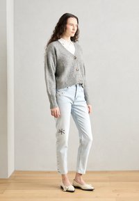 Grå ribbestrikket cardigan med treknapper, iført over en hvit skjorte med krage. Kombinert med lyseblå jeans med blomsterbroderi og beige sko.