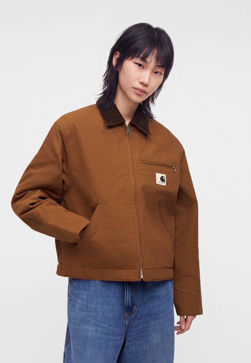 Person wearing a brown zip-up jacket with a dark collar and a small logo patch, paired with loose blue jeans, standing against a plain background.