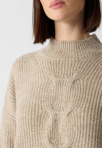 Beżowy sweter wykonany na szydełku z wysokim, prążkowanym dekoltem, wzorami o fakturze i luźnym kroju. Miękka mieszanka wełny.