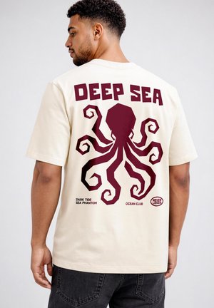 Mann trägt cremefarbenes T-Shirt mit großem stilisiertem dunkelrotem Oktopus und den Schriftzügen "DEEP SEA", "DARK TIDE SEA PHANTOM", "OCEAN CLUB" sowie einem "NEVERLESS"-Logo.