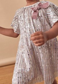 Silvrig paljetterklänning med korta fjäderlika ärmar, prydd med en ljusrosa sløjfe på axeln. Randig mönster med varierande färger och texturer.
