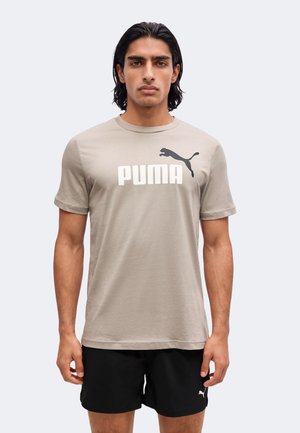 Homme aux cheveux foncés portant un t-shirt beige Puma et un short noir Puma, debout devant un fond clair uni.