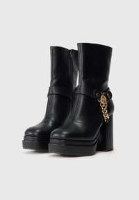 Fekete, magas sarkú, platform talpú bokacsizmák oldalsó cipzárral, díszes pánttal és arany lánc részletekkel egy egyszerű háttér előtt.