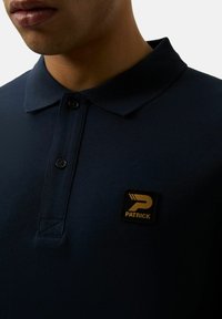 Dunkelblaues Poloshirt mit Kragen, zwei Knöpfen und einem schwarzen quadratischen Patch mit goldenem "PATRICK"-Logo. Weicher Stoff, lässiges Design.