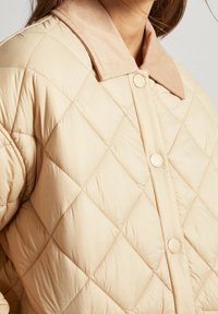 Chaqueta acolchada beige con cuello de pana, con cierre de botones y textura de patrón de diamantes. Tejido suave y ligero.