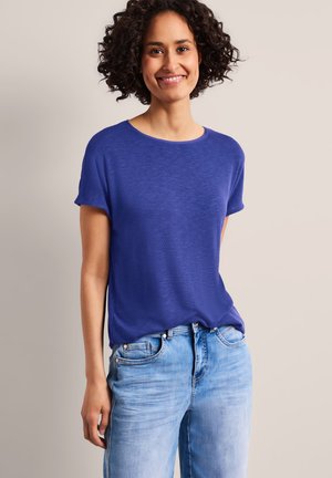 Kobieta z kręconymi włosami uśmiechająca się, ubrana w niebieską krótką koszulkę i jasnoniebieskie dżinsy, stojąca na jednolitym beżowym tle.