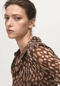 Mujer de piel clara y cabello corto castaño, lleva una blusa marrón con estampado de leopardo, mirando por encima del hombro contra un fondo liso.