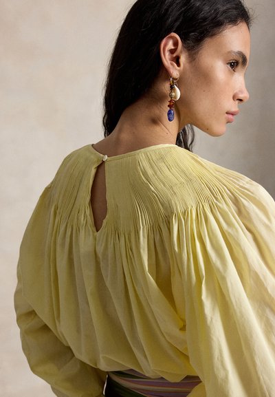 Mujer con blusa amarilla plisada de mangas largas, mostrando detalle de botones en la espalda, con pendientes colgantes de piedras multicolores, mirando hacia la derecha.