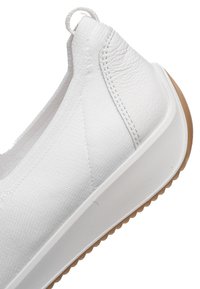 White slip-on sneaker with textured fabric upper, leather heel patch, loop pull tab, thick white sole, and brown rubber outsole.