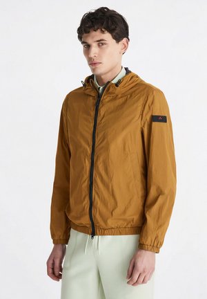 Giovane uomo che indossa una giacca con cappuccio giallo senape con zip, polsini elastici e pantaloni bianchi su uno sfondo bianco uniforme.