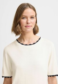 Creme kortærmet sweater med en struktureret, bølget sort halsudskæring og ærmer. Materialet ser blødt og let tekstureret ud.