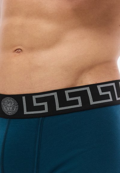 Κοντινό πλάνο γυμνού κορμού με μπλε εσώρουχο που έχει μαύρη μέση με γκρι γεωμετρικό σχέδιο και έμβλημα κεφαλής Μέδουσας.