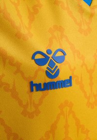 Gul texturerad sporttröja med en blå "hummel"-logotyp och emblem, med ett distinkt mönster.