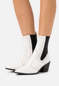 Botas de tobillo de imitación cuero blancas con lados elásticos negros, detalles de tachuelas, punteras puntiagudas y un tacón corto y negro.