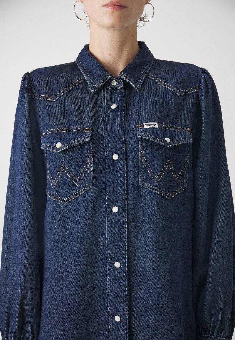 Chemise en jean bleu foncé avec un col classique, des manches bouffantes, deux poches poitrine et des coutures en zigzag contrastées. Équipée de boutons pression blancs.