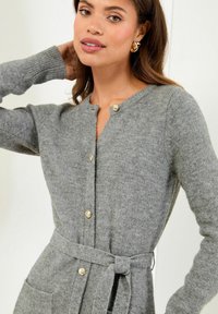 Frau in einer grauen gestrickten Strickjacke mit goldenen Knöpfen und einem gebundenen Gürtel, die mit einer Hand hinter ihrem Kopf vor einem schlichten Hintergrund posiert.