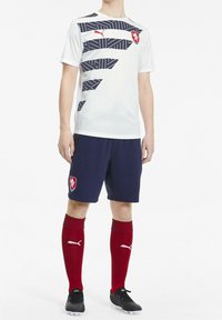 Bílý fotbalový dres s námořnickými geometrickými vzory, doplněný námořnickými šortkami, červenými ponožkami a černými kopačkami. Obsahuje logo na hrudi a na šortkách.