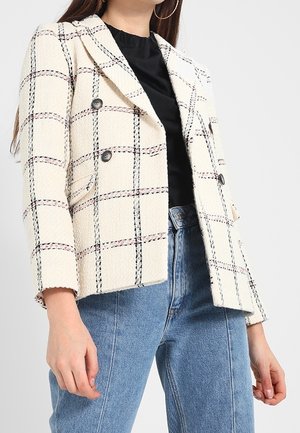 Mujer vestida con un blazer a cuadros color crema de doble botonadura sobre una blusa negra y jeans azul de talle alto, mostrada desde el cuello hasta las caderas.