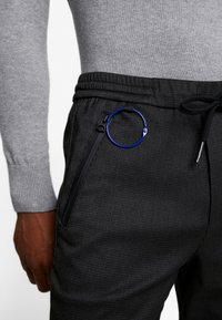 Czarne joggery o fakturowanej powierzchni, z kieszenią na zamek i niebieskim metalowym kółkiem jako ozdobą. Talia jest elastyczna, z regulacją za pomocą sznurka.