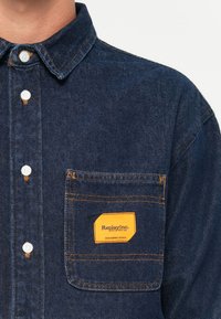 Dark blue denim shirt featuring a buttoned collar, white buttons, and an orange label on the chest pocket with "Replay Inc." text.