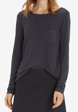 Femme portant une chemise sombre à manches longues avec une petite poche sur la poitrine gauche, rentrée dans une jupe sombre.