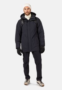 Chaqueta acolchada azul marino con capucha, cierre de cremallera y múltiples bolsillos. Combinada con pantalones oscuros y guantes negros. Gorro de punto beige en la cabeza.