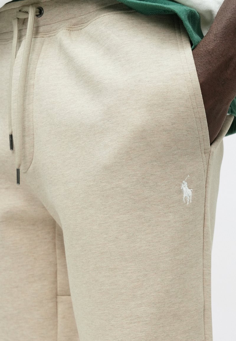 Beige joggers met een zachte textuur, trekkoordsluiting en zijzakken. Heeft een klein wit geborduurd logo op de dij.