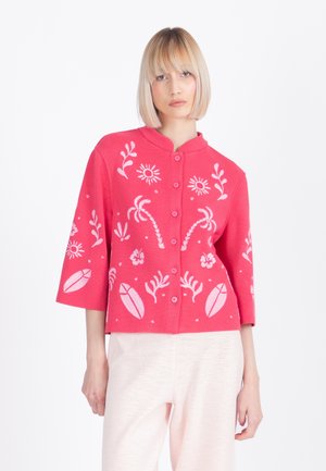 Kobieta ubrana w koralowo-różowy kardigan na guziki z białymi tropikalnymi liśćmi i wzorami słońca, w połączeniu z jasnobeżowymi spodniami.