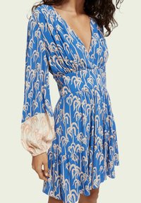 Modrá šaty s květinovým vzorem v světle růžové barvě, s hlubokým výstřihem do V, dlouhými puff rukávy a volánkovým lemem. Vyrobeno z texturované tkaniny.
