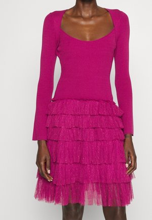 Kobieta w jasnoróżowej prążkowanej bluzce z długim rękawem, połączonej z warstwową tiulową spódnicą z falbanami w tym samym odcieniu różu.