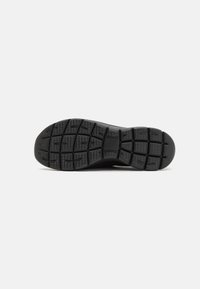 Suela negra de zapatilla con una superficie de goma texturada; presenta un patrón en la suela para mejorar la tracción y estabilidad; punta redondeada y talón pronunciado.