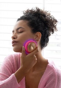 Round, pink facial cleansing device with a textured gold surface held against cheek, glowing blue light, soft pink sweater, natural light setting.