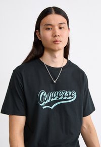 Črna bombažna majica s širokim, svetlo modrim logotipom "Converse". Model nosi srebrno verižico. Preprost, sproščen dizajn.
